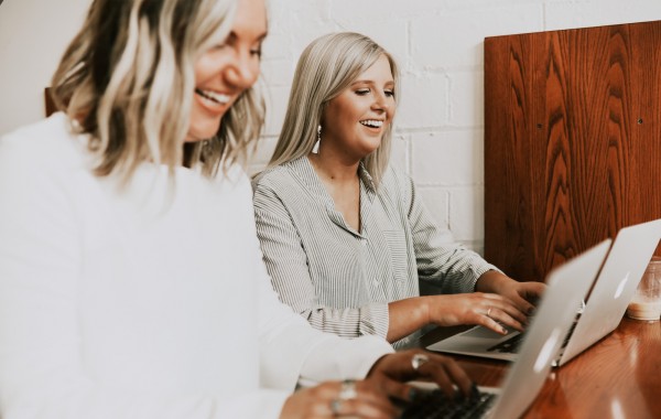Two laughing employees are working side by side on their modern laptops.