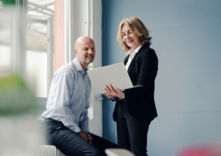 Two employees wearing business outfits are looking at a laptop screen and discussing their work.