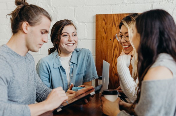 A group of young people in casual clothes is standing around a table discussing their work.