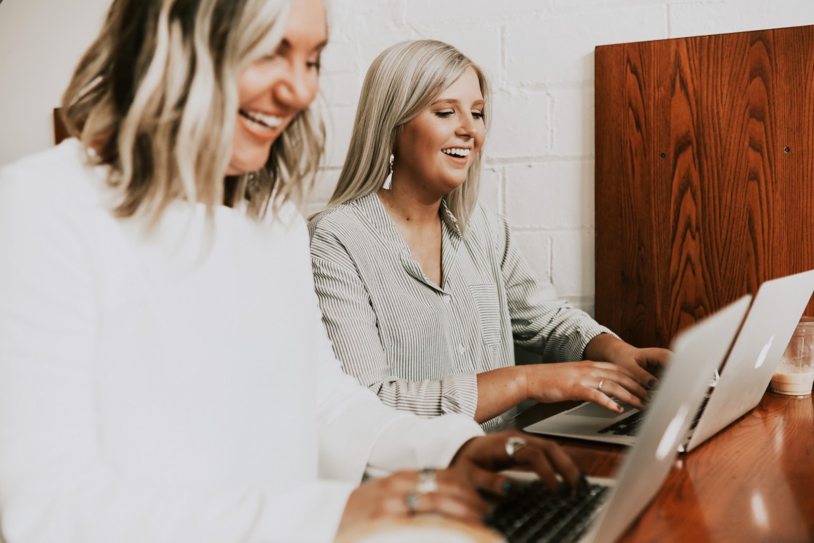 Two laughing employees are working side by side on their modern laptops.