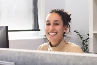 Nahaufnahme einer jungen Mitarbeitenden, die hinter einer Hotelrezeption sitzt und direkt in die Kamera lacht.