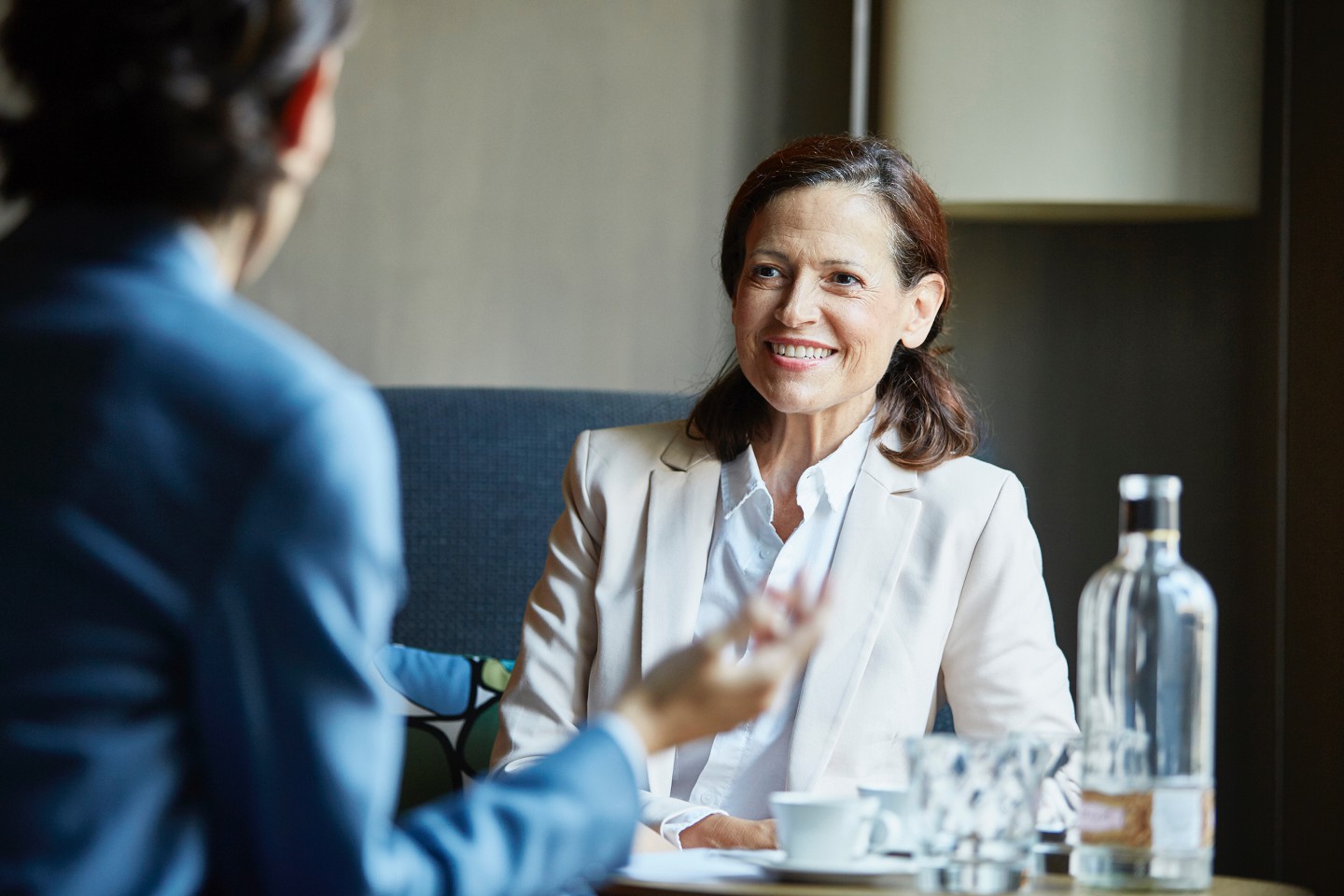 Two people are sitting at a small coffee table, talking to each other. They wear business outfits.